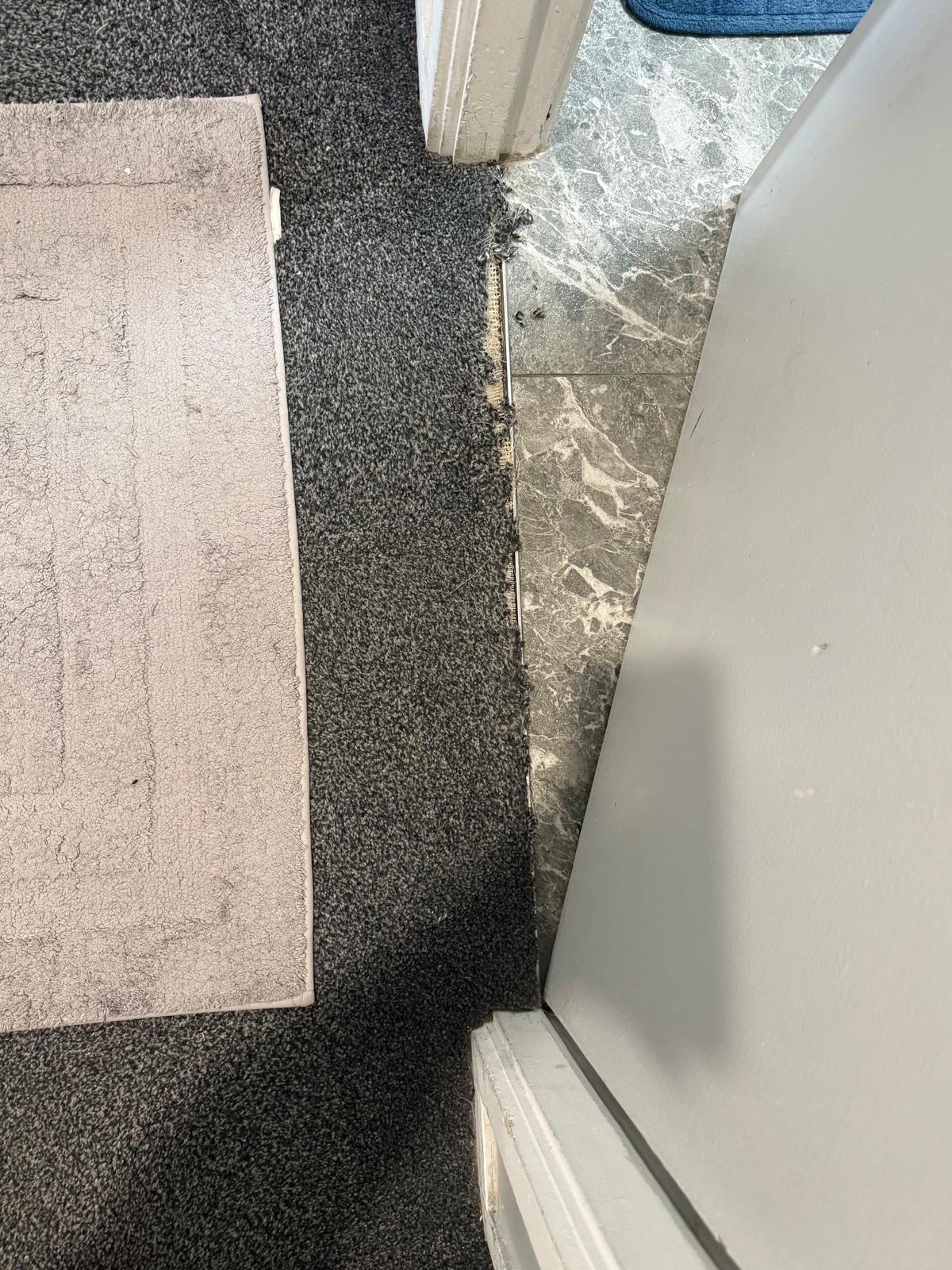 Charcoal carpet frayed and pulling away from the gripper at a bathroom doorway beside grey marble tile